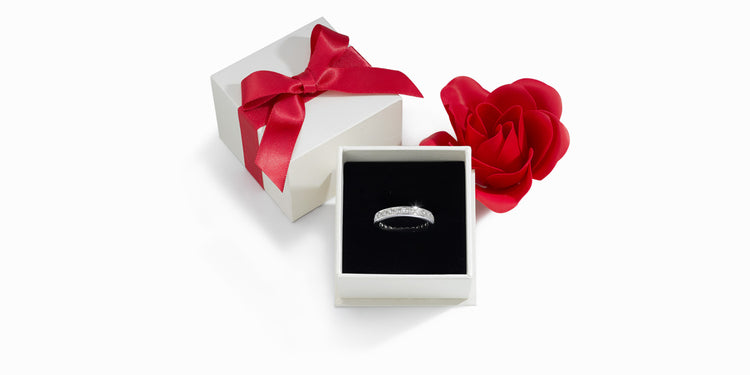 Eleganter Diamantring in weißer Schmuckschachtel mit schwarzem Samt-Inlay, daneben weiße Geschenkbox mit roter Satinschleife und rote Rosenblüte auf weißem Hintergrund