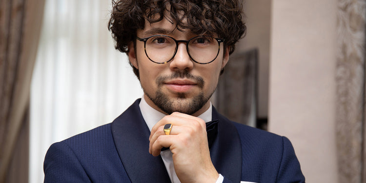 Eleganter Mann mit lockigen Haaren und Brille in dunkelblauem Anzug trägt goldenen Siegelring
