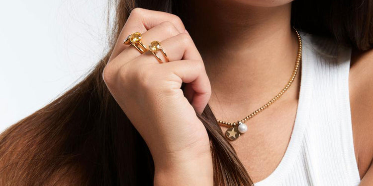 Frau mit langen braunen Haaren in weißem Top trägt mehrere goldene Ringe mit Kugel-Design und goldene Halskette mit Stern- und Perlenanhänger 